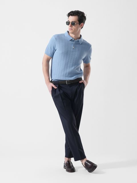 Uomo con occhiali da sole, che indossa una polo a maniche corte azzurra, pantaloni scuri e mocassini marroni, in piedi con un piede incrociato davanti all'altro.