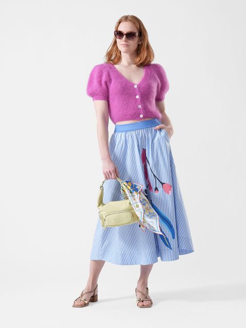 Donna con un top rosa peloso, una gonna midi blu a righe con motivo floreale, occhiali da sole, una borsa gialla con foulard e sandali.
