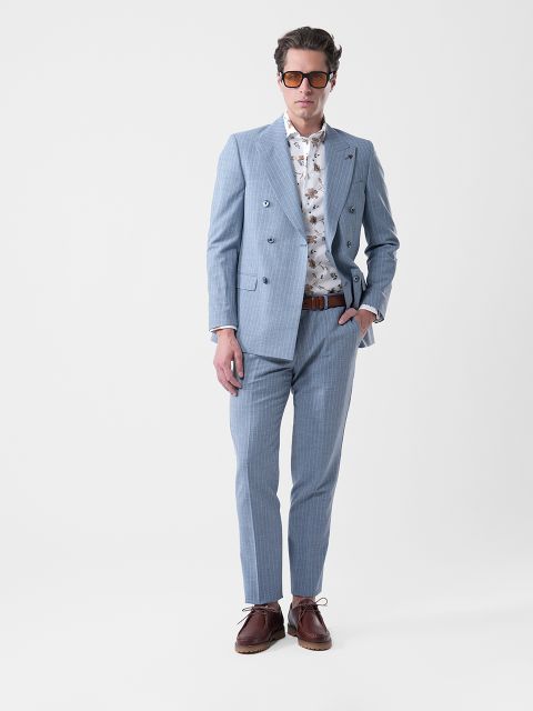 Uomo in abito azzurro, camicia bianca a fantasia, scarpe marroni e occhiali da sole, in piedi con una mano in tasca davanti a uno sfondo semplice.