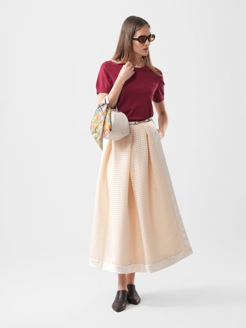 Donna con top rosso, gonna lunga color crema, scarpe nere, occhiali da sole e una borsa a fantasia in mano, in piedi davanti a uno sfondo semplice.