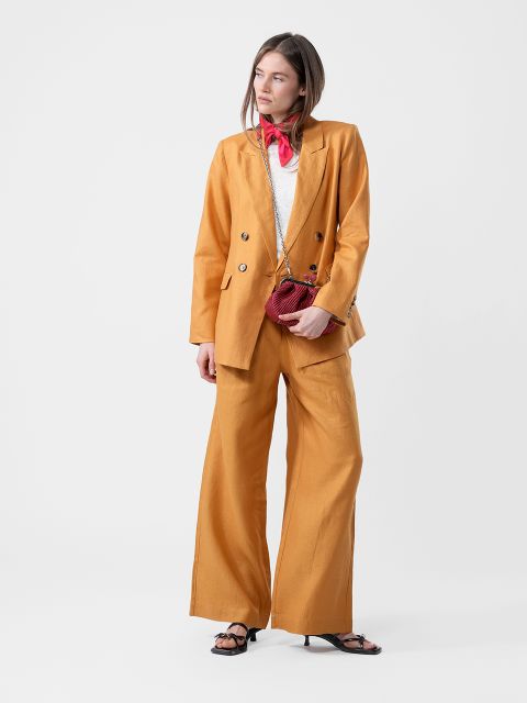 Donna in completo giallo senape, top bianco, sciarpa rossa, con una piccola borsa rossa e sandali neri, davanti a uno sfondo bianco semplice.