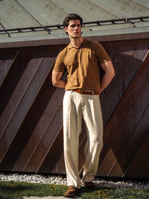 Un uomo è in piedi all'aperto, indossa una polo marrone, pantaloni a pieghe color crema e scarpe marroni ed è appoggiato a un edificio di metallo alla luce del sole.