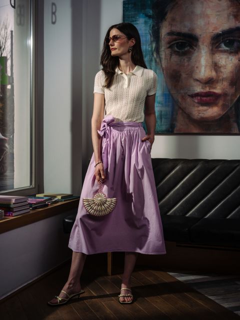 Una donna indossa una polo color crema e una gonna fluida color lavanda e tiene in mano una borsa a forma di ventaglio decorata con perline.