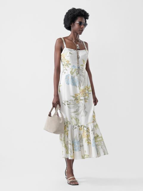 Una donna in un abito da sole bianco a fiori, sandali, occhiali da sole e collana, che indossa una borsetta beige, posa davanti a uno sfondo chiaro e semplice.