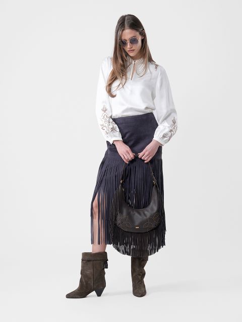 Donna con occhiali da sole, camicetta bianca, gonna nera con frange, stivali marroni e una borsa nera in mano, davanti a uno sfondo semplice.