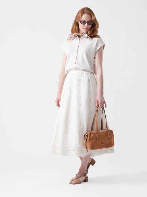 Donna con occhiali da sole, camicetta bianca e gonna lunga bianca, che tiene una borsa color marrone chiaro e indossa sandali beige, in piedi davanti a uno sfondo bianco.