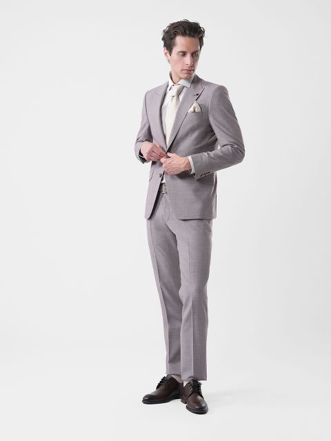 Uomo in abito grigio chiaro, camicia color crema e cravatta, in piedi davanti a uno sfondo bianco semplice, che si abbottona la giacca e guarda di lato.