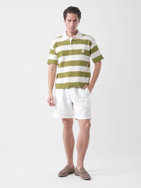 Uomo in polo a righe bianche e verdi, pantaloncini bianchi e scarpe basse marroni davanti a uno sfondo bianco semplice.