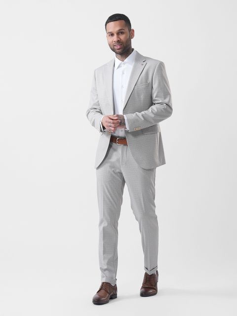 Uomo in abito grigio chiaro con camicia bianca e scarpe marroni, in posa davanti a uno sfondo bianco semplice.