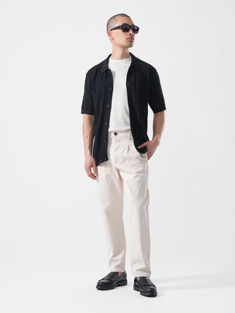 Uomo con occhiali da sole che indossa una camicia nera a maniche corte sopra una t-shirt bianca, pantaloni color crema e scarpe nere, in piedi davanti a uno sfondo bianco.