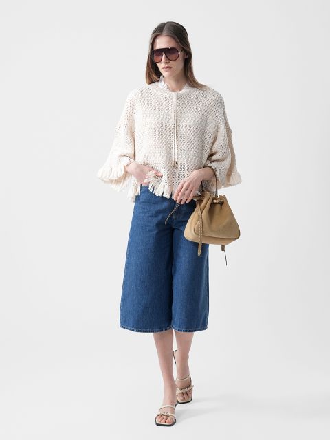 Donna che indossa occhiali da sole, un maglione di maglia color crema con frange, culottes di jeans blu al ginocchio e sandali, e tiene una borsa color marrone chiaro su uno sfondo bianco.