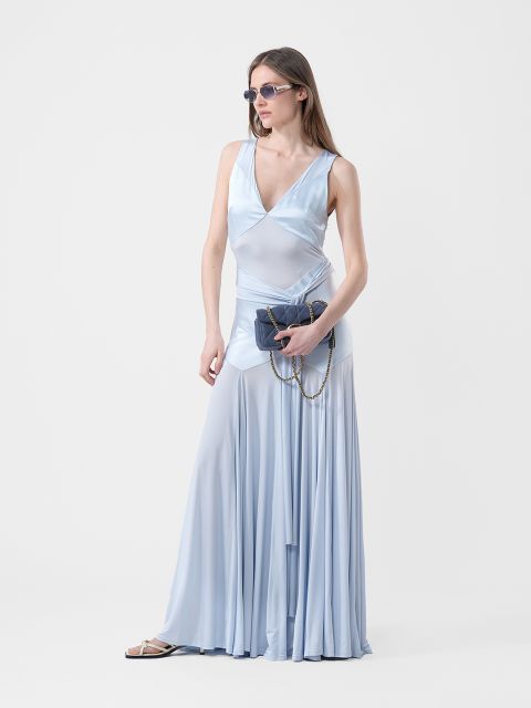 Donna in un abito azzurro chiaro, fluido e senza maniche, con occhiali da sole, che tiene in mano una borsa scura con catena, in piedi davanti a uno sfondo bianco semplice.