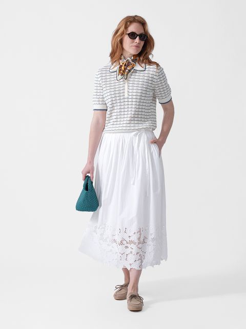 Una donna indossa occhiali da sole, un top a righe, una gonna midi bianca con ricami, una sciarpa al collo, una borsa verde acqua e scarpe beige.