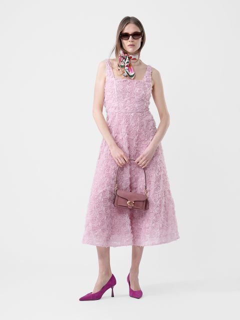 Donna in un vestito rosa testurizzato senza maniche, tacchi viola, occhiali da sole, una sciarpa al collo e con una borsa viola in mano, in piedi davanti a uno sfondo bianco.