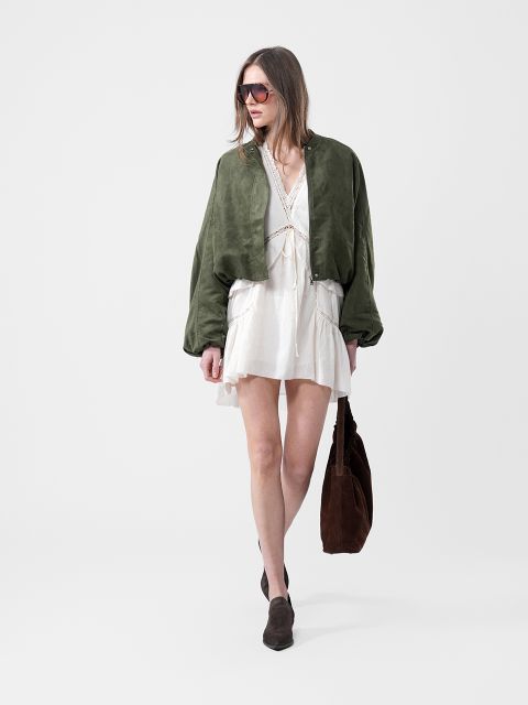 Donna in abito bianco, giacca verde, occhiali da sole e stivali marroni che porta una grande borsa marrone davanti a uno sfondo bianco semplice.