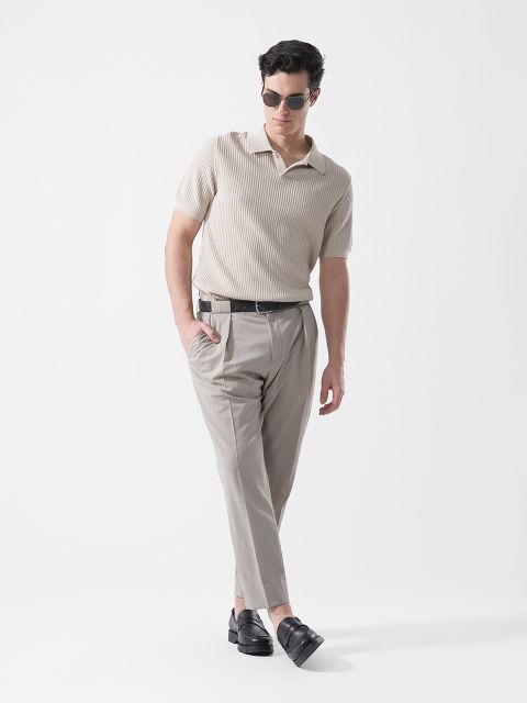 Un uomo che indossa occhiali da sole, una camicia in maglia beige a maniche corte, pantaloni grigio chiaro e mocassini neri, in piedi davanti a uno sfondo bianco semplice.