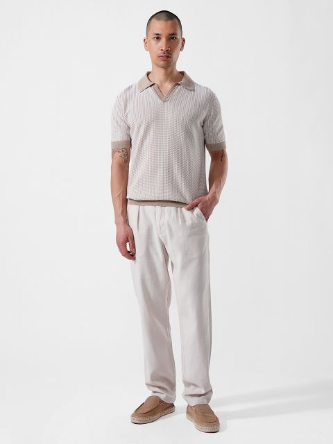 Un uomo è in piedi davanti a uno sfondo bianco e indossa una camicia beige a maniche corte con colletto, pantaloni chiari e mocassini marrone chiaro.