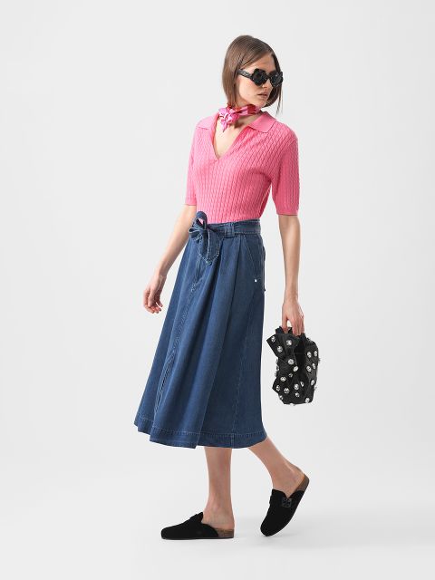 Frau mit rosa Oberteil, blauem Midirock, schwarzen Halbschuhen, Sonnenbrille und rosa Halstuch, hält eine schwarze Blumentasche und schaut zur Seite.