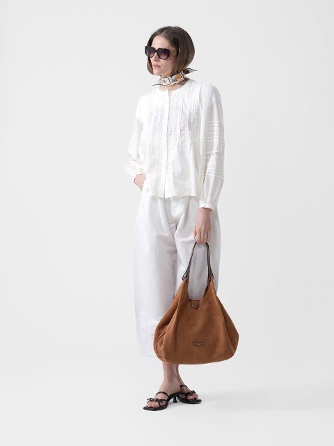 Donna con occhiali da sole, camicetta e pantaloni bianchi, una sciarpa al collo, sandali neri e una grande borsa a mano in pelle scamosciata marrone.