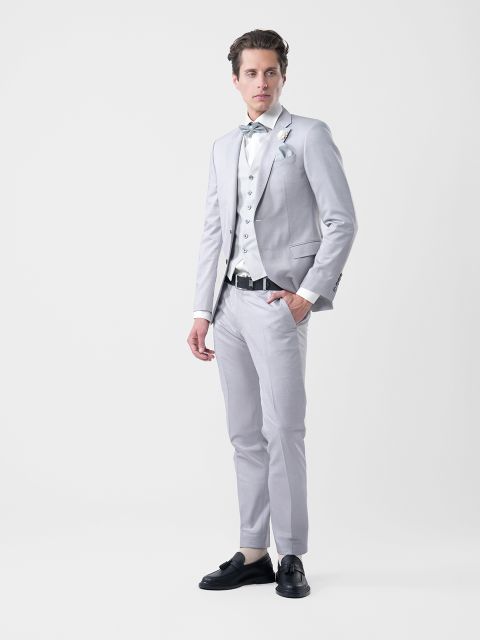 Uomo in abito grigio chiaro con camicia bianca, gilet fantasia e papillon, in piedi davanti a un semplice sfondo bianco.