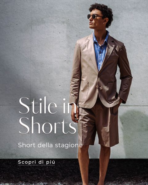 Un uomo con occhiali da sole indossa una giacca da completo marrone con pantaloncini abbinati e una camicia a righe blu. Un testo italiano sullo stile dei pantaloncini estivi è sovrapposto.