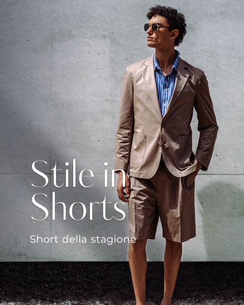 Un uomo con occhiali da sole indossa una giacca da completo marrone chiaro con pantaloncini abbinati e una camicia a righe blu. Il testo recita "Stile in Shorts-Short della stagione."