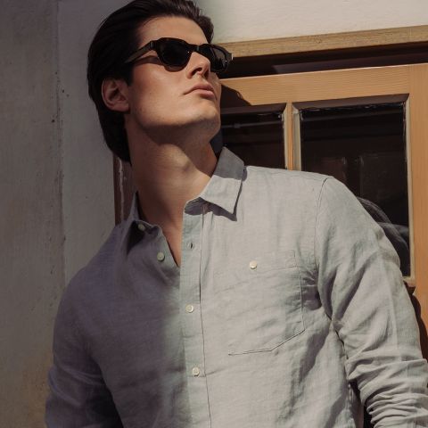 Un uomo con occhiali da sole e una camicia button-up grigio chiaro è in piedi alla finestra e guarda il sole.