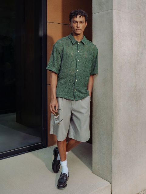 Un uomo con una camicia verde a maniche corte, pantaloncini grigi, scarpe basse nere e calzini bianchi sta in piedi accanto a un muro di cemento e indossa occhiali da sole.