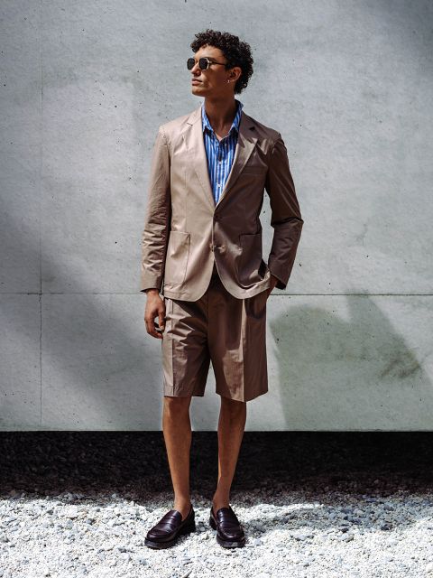 Un uomo che indossa occhiali da sole, un blazer marrone chiaro con pantaloncini abbinati, una camicia a righe blu e scarpe basse, è in piedi davanti a un muro di cemento su ghiaia.