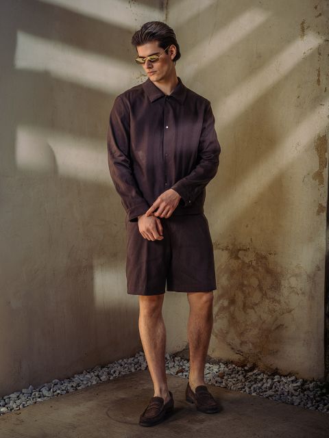 Un uomo con occhiali da sole, una camicia abbottonata marrone scuro, pantaloncini abbinati e scarpe basse sta in piedi davanti a un muro strutturato e illuminato dal sole con accenti di pietra.