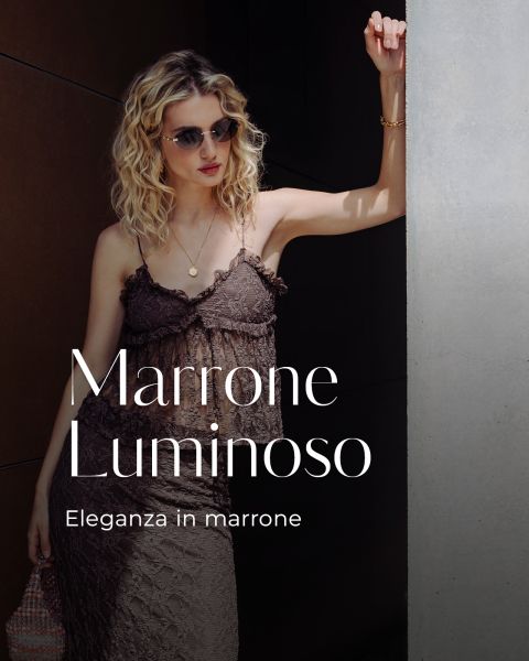 Donna bionda con occhiali da sole indossa un vestito di pizzo marrone e sta in piedi accanto a un muro. Il testo recita "Marrone Luminoso. Eleganza in marrone."