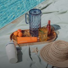Un vassoio a bordo piscina con bicchieri, albicocche, crema solare, uno spray, un cappello da sole, una tazza bianca e una stella marina su un asciugamano verde chiaro.