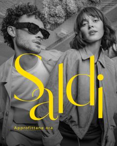 Un uomo e una donna eleganti posano insieme in outfit alla moda. Il testo giallo recita "Saldi" e "Approfittane ora" e pubblicizza una vendita.