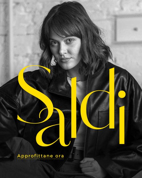Una donna in giacca di pelle siede con sicurezza; in grande scritta gialla c'è scritto "Saldi" e in una scritta più piccola "Approfittane ora".