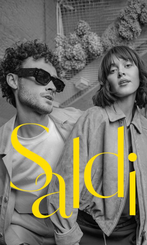 Un uomo elegante e una donna elegante posano in giacche e occhiali da sole; il testo giallo e in grassetto "Saldi" è sovrapposto alla foto in bianco e nero.