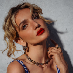 Una donna con capelli biondi mossi, rossetto rosso audace e gioielli d'oro posa davanti a un muro di cemento, toccandosi il collo e guardando con sicurezza la fotocamera.