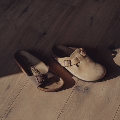Due sandali Birkenstock spaiati - uno a punta aperta e uno a punta chiusa - con fibbie su un pavimento di legno illuminato dal sole.