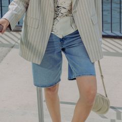Una persona che indossa un blazer a righe chiare, una blusa stampata e shorts di jeans è in piedi all'aperto vicino a una ringhiera, tenendo in mano una piccola borsetta beige.