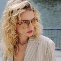 Una donna con capelli biondi ricci, che indossa occhiali da sole aviator colorati, un blazer beige a righe e una collana a catena spessa, è in piedi vicino a uno specchio d'acqua.