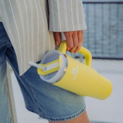 Persona in pantaloncini di jeans e giacca a righe che tiene una grande tazza Stanley gialla con cannuccia e manico.