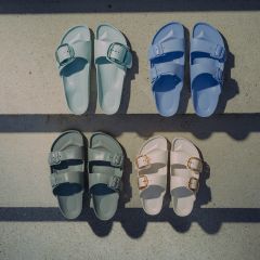 Quattro paia di sandali in blu pastello, verde pastello, verde scuro e bianco sporco, disposti su una superficie di cemento e attraversati da ombre.