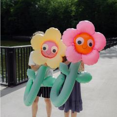 Due persone si tengono grandi fiori gonfiabili gialli e rosa davanti al viso, mentre stanno su un sentiero soleggiato vicino a una ringhiera e a delle piante verdi.