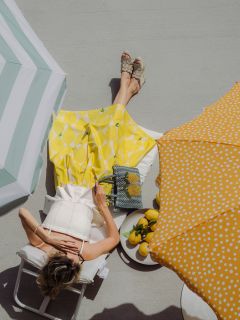 Una donna in gonna gialla si rilassa su una sedia a sdraio sotto gli ombrelloni in una giornata di sole, con limoni e una borsa a fantasia di limoni accanto a sé.