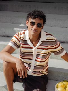 Un uomo con i capelli ricci e gli occhiali da sole è seduto su una scalinata esterna e indossa una camicia a righe a maniche corte; accanto a lui c'è una ciotola con dei limoni.