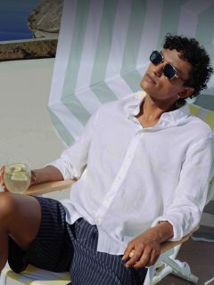 Un uomo con occhiali da sole e una camicia bianca si rilassa su una sedia a sdraio, con in mano una bevanda al limone, sotto un ombrellone a strisce in una giornata di sole.