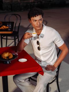 Uomo in camicia bianca seduto a un tavolo rosso, beve un espresso, ha un croissant e un bicchiere d'acqua davanti a sé in un caffè all'aperto.