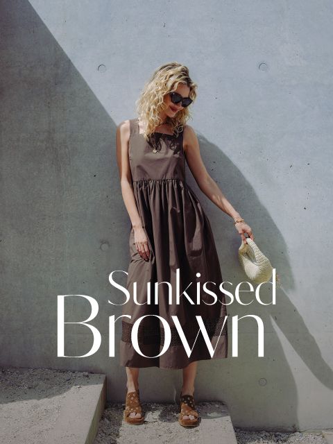 Una donna in un vestito marrone senza maniche sta in piedi accanto a un muro di cemento e tiene in mano una borsa di paglia. Il testo recita "Sunkissed Brown".