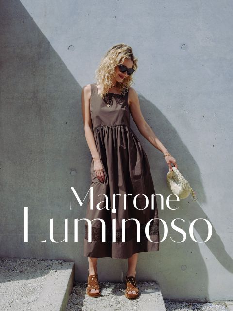 Una donna in un vestito marrone senza maniche e sandali si appoggia a un muro di cemento e tiene in mano un cappello di paglia. Il testo recita: „Marrone Luminoso“.