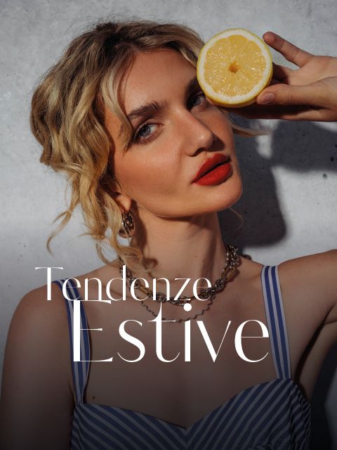 Una donna con capelli biondi ondulati tiene mezzo limone davanti al viso; indossa un top a righe e rossetto rosso. Il testo recita: "Tendenze Estive".