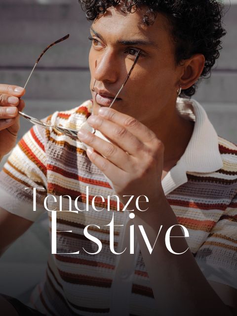 Un giovane uomo con i capelli ricci, che tiene in mano degli occhiali da sole, indossa una camicia a maglia a righe e siede all'aperto. Il testo recita: „Tendenze Estive.“